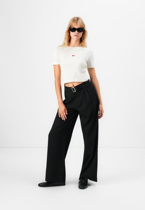 Woman wearing black sunglasses, white cropped t-shirt with heart logos, black wide-leg trousers with belt, and black shoes, standing against white background.