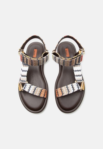 Ζευγάρι καφέ επίπεδες σανδάλες με πολύχρωμες υφασμάτινες λουρίδες ζιγκ-ζαγκ και κλεισίματα στον αστράγαλο, εμφανίζονται από πάνω σε απλό φόντο.
