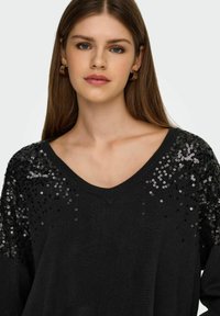 Pull noir avec un décolleté en V et des manches longues, orné de sequins noirs et argentés sur les épaules. Matière en tricot douce.