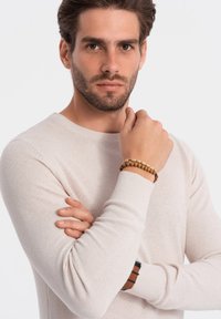 Pull en maille texturée beige clair avec des manches longues et un col rond. Le modèle porte un bracelet en perles de bois à un poignet.