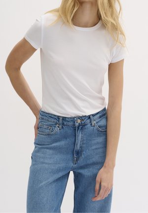 Blonde woman wearing a plain white short-sleeve t-shirt tucked into blue high-waisted jeans against a plain background.