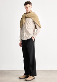 Béžová pruhovaná košeľa, čierne nohavice a hnedé semišové členkové čižmy. Vlnený sveter prehodený cez ramená. Minimalistický dizajn, neutrálne farby.