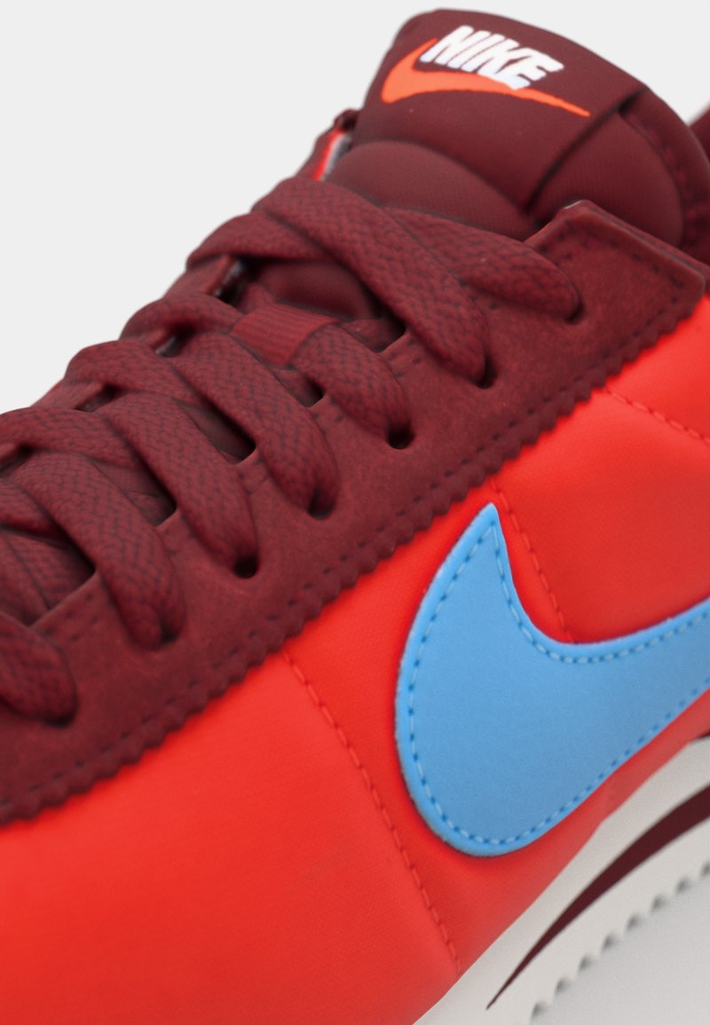 Zapatilla roja y naranja con parte superior texturizada, swoosh azul de Nike y cordones a juego. Cuenta con una suela de goma y un cuello acolchado.