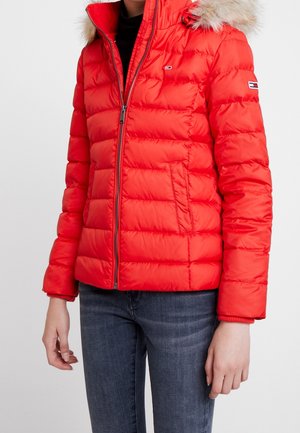 Rote Daunenjacke mit einer Kapuze aus Kunstpelzbesatz, Reißverschluss vorne, gesteppten Paneelen und elastischen Bündchen. Kombiniert mit dunklen Jeans.