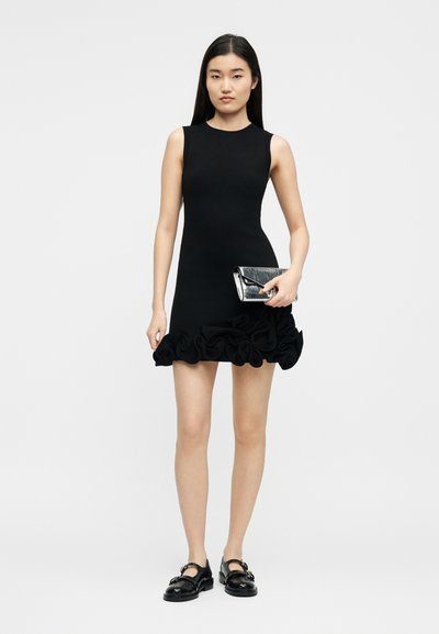 Γυναίκα που φοράει μαύρο φόρεμα χωρίς μανίκια με βολάν στο τελείωμα, κρατώντας ένα μικρό ασημένιο τσαντάκι και φοράει μαύρα παπούτσια Mary Jane, στέκεται σε λευκό φόντο.