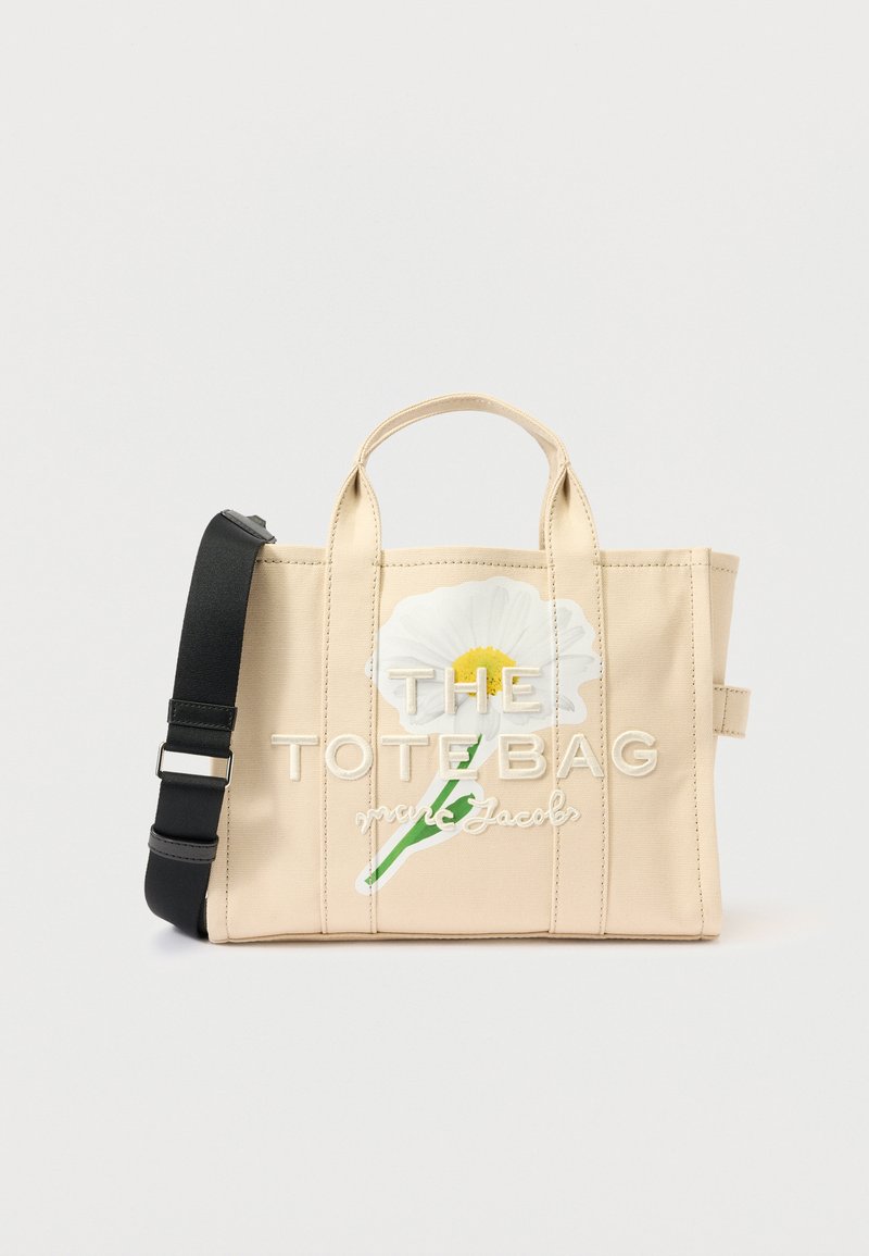 Béžová plátenná taška s dvoma hornými uchami a odnímateľným čiernym popruhom. Obsahuje výšivku bielych kvetov a názov značky písmom tučným písmom.