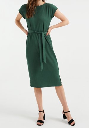 Femme portant une robe verte mi-longue à manches courtes et ceinture nouée à la taille, accompagnée de sandales noires à talons.