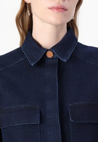 Giacca di jeans blu scuro con colletto classico, due tasche frontali e un distintivo dettaglio con bottone al collo. Texture liscia.