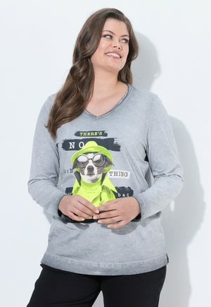 Kvinde smilende iført en grå langærmet skjorte med en hundeillustration i neonjakke og tekst foran, stående mod en hvid væg.