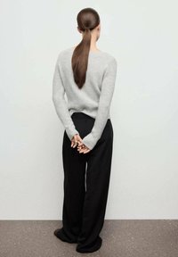 Suéter gris de punto con un escote en la espalda, combinado con pantalones de pierna ancha negros. Cabello largo recogido. De pie frente a un fondo neutro.