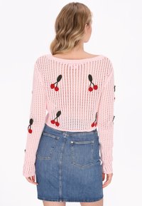 Suéter de punto rosa claro con un patrón de agujeros abiertos, que presenta bordados de cerezas rojas. Combinado con una falda corta de mezclilla, que muestra bolsillos.
