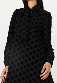 Robe noire à pois avec un col à nœud, présentant une texture transparente et des manches bouffantes. Les pois sont en relief, créant un effet tridimensionnel.