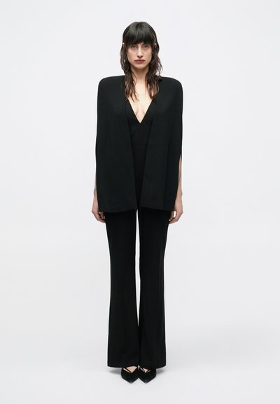 Combinaison noire avec un décolleté en V profond et un design à jambes larges, surmontée d'une cape noire structurée dotée d'ouvertures au niveau des bras.
