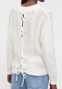 Personne portant un pull en maille torsadée blanche avec un détail lacé dans le dos et un jean denim bleu clair, vue de dos.