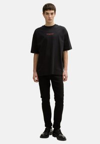 Black oversized t-shirt with short sleeves, featuring red text on the chest, paired with black slim-fit trousers and black lace-up shoes.