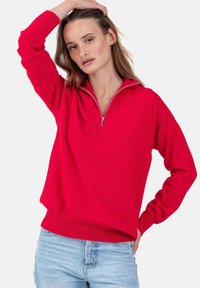 Pull rouge zippé en tissu doux, avec manches longues et coupe décontractée. Porté avec un jean en denim bleu clair.