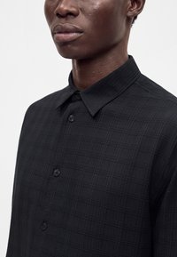 Gros plan d'une personne portant une chemise noire à carreaux avec col et boutons, montrant le cou et le bas du visage sur un fond uni.