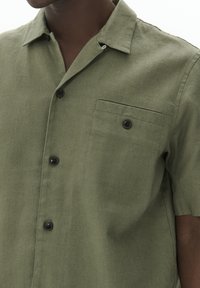 Camicia a maniche corte in tessuto verde oliva con una finitura testurizzata. Presenta una tasca sul petto e bottoni neri lungo l'apertura frontale.