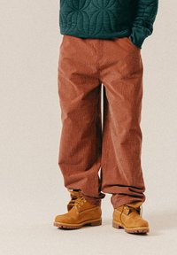 Braun karierte Hose in lockerer Passform, hergestellt aus strukturiertem Stoff, kombiniert mit hellbraunen Wildlederstiefeln mit gelben Schnürsenkeln und einer robusten Sohle.