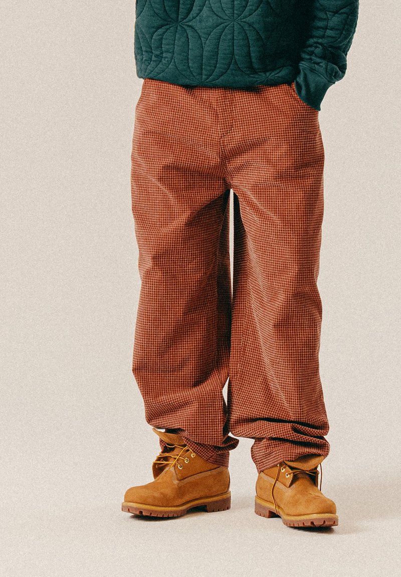 Braun karierte Hose in lockerer Passform, hergestellt aus strukturiertem Stoff, kombiniert mit hellbraunen Wildlederstiefeln mit gelben Schnürsenkeln und einer robusten Sohle.