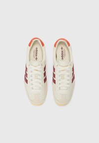 Vita och mörkröda Adidas-sneakers med perforerad textur, med tre mörkröda ränder, krämfärgade skosnören och orange accenter på hälen.