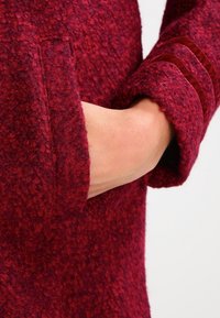 Rode, gestructureerde coat met een zachte, pluizige afwerking; heeft een omgeslagen manchet en een zijzak, die warmte en functionaliteit benadrukken.