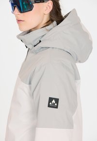 Grå og hvid jakke med hætte, som har en blød tekstur, en sort logo-patch på ærmet og et elegant design.