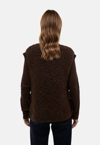 Gilet sans manches marron en laine douce, avec un dos texturé, un haut à manches longues marron clair en dessous, et un jean bleu foncé.