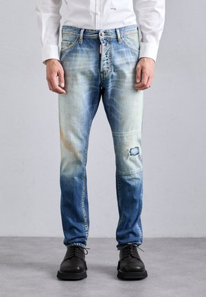 Uomo che indossa jeans blu sbiaditi con un strappo sul ginocchio destro, scarpe nere con lacci e una camicia bianca a maniche lunghe, in piedi su un pavimento grigio.