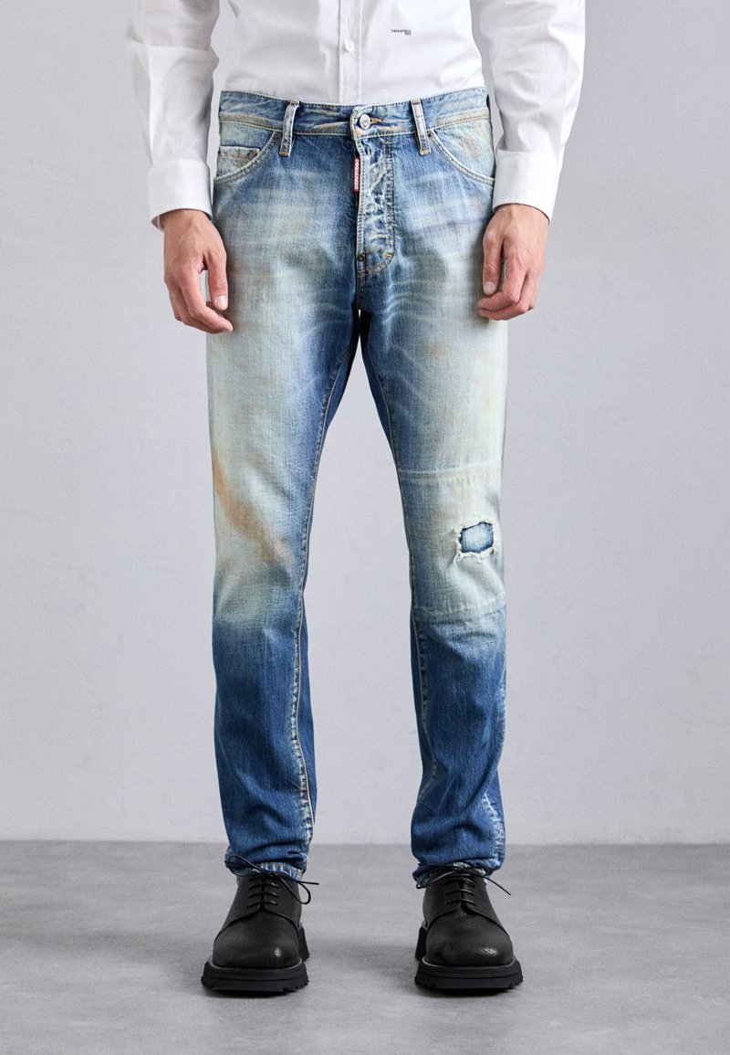 Uomo che indossa jeans blu sbiaditi con un strappo sul ginocchio destro, scarpe nere con lacci e una camicia bianca a maniche lunghe, in piedi su un pavimento grigio.