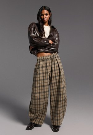 Femme debout, les bras croisés, portant une veste en cuir marron, un haut crème, un pantalon large à carreaux et des chaussures foncées, sur un fond gris.