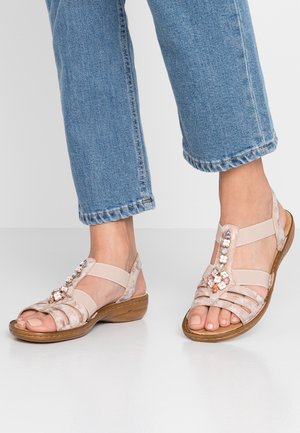 Pieds chaussés d’un jean bleu et de sandales beige à bout ouvert avec des ornements floraux et des brides élastiques, debout sur un sol blanc.