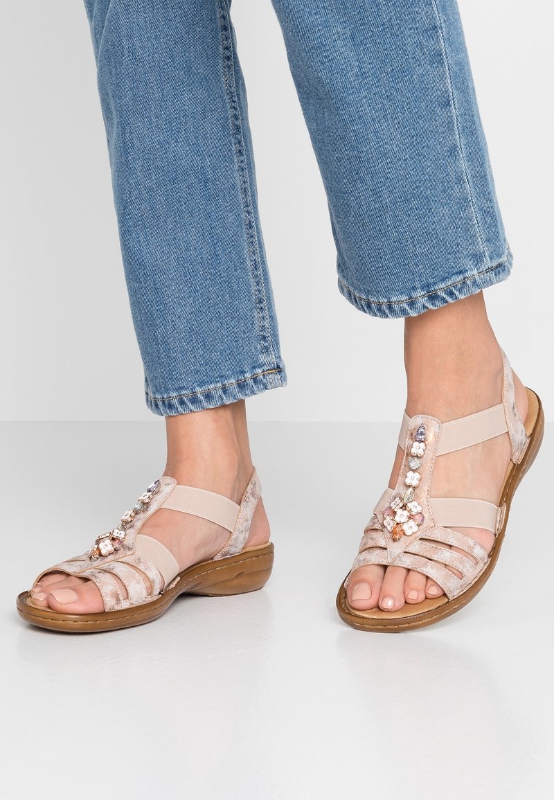 Pieds chaussés d’un jean bleu et de sandales beige à bout ouvert avec des ornements floraux et des brides élastiques, debout sur un sol blanc.