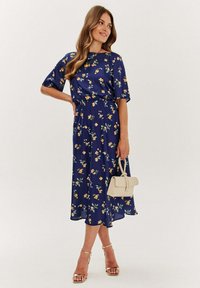 Granatowa w kwiaty midi sukienka z krótkimi rękawami, z żółtymi i zielonymi wzorami. Noszona z metalicznymi szpilkami i trzymana w małej beżowej torebce.