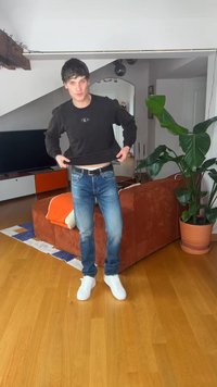 Νεαρός άνδρας με μαύρο φούτερ και τζιν σηκώνει ελαφρώς το πουκάμισο σε ένα σαλόνι με πορτοκαλί καναπέ, μεγάλο φυτό και ξύλινο πάτωμα.