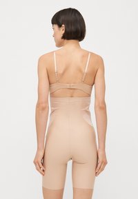 Beige heldrakt med et innsvingt design, justerbare stropper og en jevn tekstur. Har midt-lår lengde og lav scoop rygg.