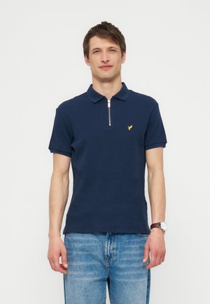 Mężczyzna noszący granatowe polo z krótkim rękawem, przednim zamkiem błyskawicznym i małym żółtym logo, w połączeniu z jasnoniebieskimi jeansami i zegarkiem na rękę.