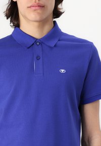 Blå polo shirt lavet af struktureret bomuldsstof. Har en klassisk krave, tre-knaps knaplukning og et lille logo på brystet.