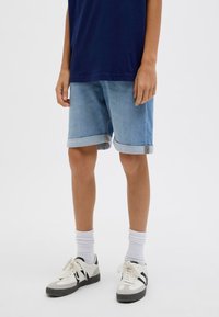 Shorts in denim di colore azzurro chiaro con risvolti, indossati con una maglietta scura, calze bianche e sneakers nere e bianche.