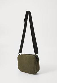 Olivengrøn corduroy crossbody-taske med rektangulær form, lynlåsclosure og justerbar sort rem. Tekstureret stof med ribbet mønster.