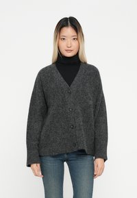 Grauer Wollcardigan mit V-Ausschnitt, Knopfverschluss und langen Ärmeln. Weiche Textur und lockere Form, getragen über einem schwarzen Rollkragenpullover.