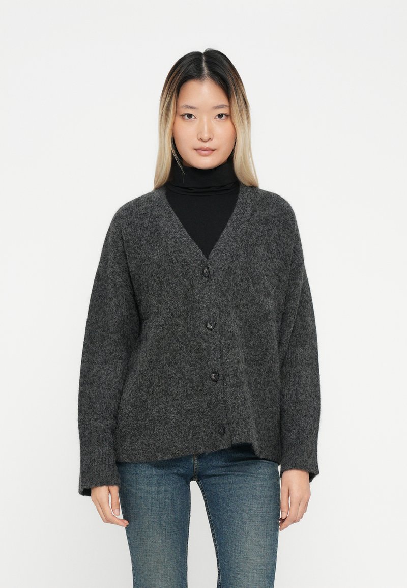 Sivi vuneni kardigan s V-izrezom, s dugim rukavima i kopčanjem sprijeda. Mekana tekstura i opušteni kroj, nosi se preko crne turtleneck majice.