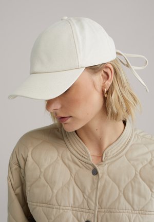 Heledate juustega naine, kes kannab beežikat tikanditega jopet ja valget pesapalli nokamütsi, millel on seotud paelad, vaadates alla ja küljele.