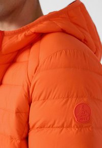 Chaqueta acolchada naranja con textura acolchada, con capucha y un parche del logo JOTT en naranja a juego en la manga.