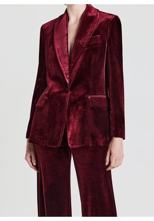 Vrouw draagt een diep rood fluwelen blazer en bijpassende broek, staande tegen een effen lichte achtergrond.