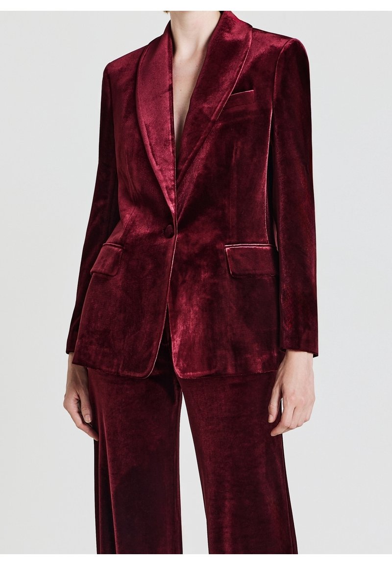 Femme portant un blazer en velours rouge profond et un pantalon assorti, debout sur un fond clair uni.