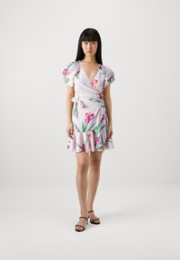 Robe portefeuille en tissu rose clair avec un imprimé floral vibrant. Présente des manches volantées, un décolleté en V plongeant et un ourlet volanté. Associée à des sandales noires.