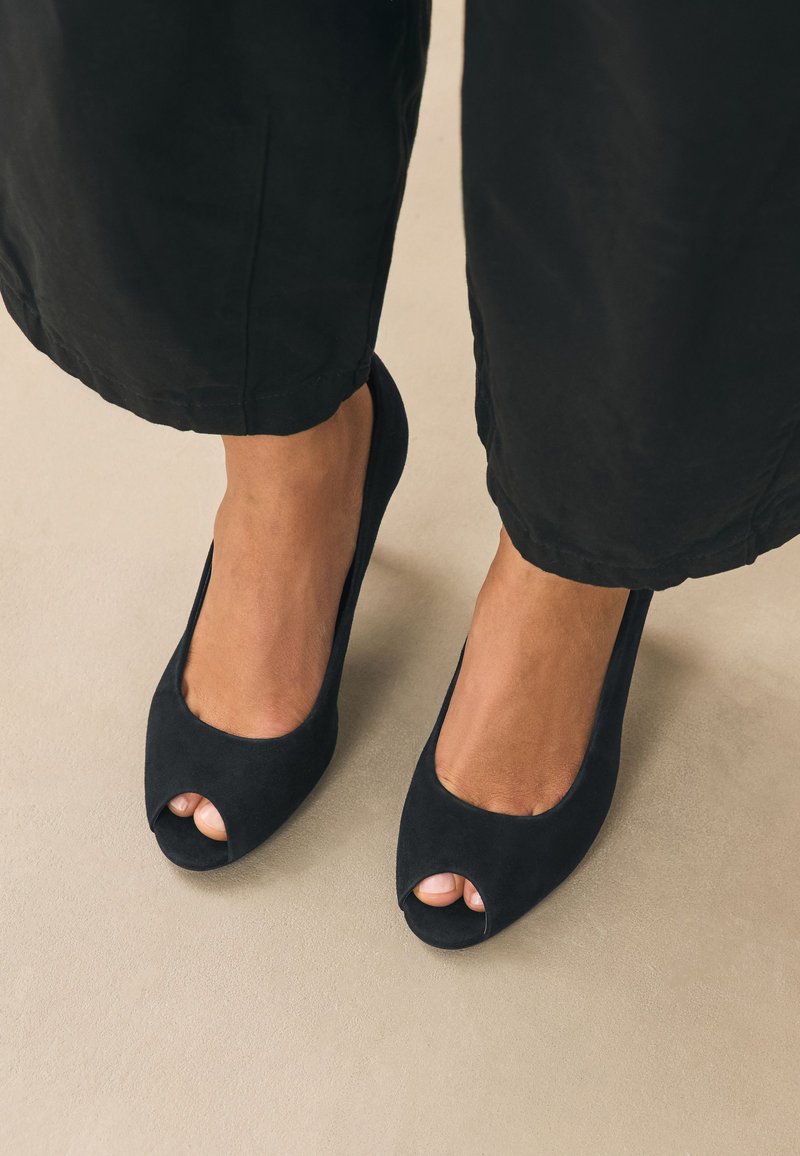 Pieds féminins portant des escarpins noirs à bout ouvert avec un pantalon noir, debout sur un sol beige.