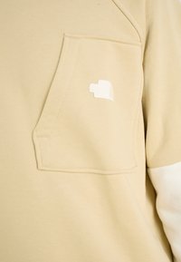 Felpa beige con una grande tasca anteriore e logo ricamato bianco. Tessuto morbido, con maniche bianche a contrasto.