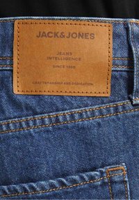 Vaqueros de mezclilla con un parche de cuero marrón. El parche presenta texto en relieve: "JACK & JONES", "JEANS INTELLIGENCE" y "CRAFTSMANSHIP AND DEDICATION".